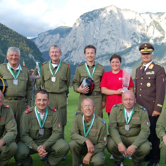 Eine Gruppe von Männern und Frauen in Militäruniformen posiert für ein Foto mit Bergen im Hintergrund. Eine Frau in einem roten Shirt hält einen Pokal.