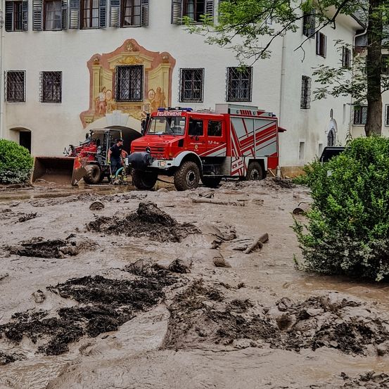 Ein roter Feuerwehrwagen und ein roter Baumaschinenwagen stecken in einem Schlammbereich vor einem Gebäude mit einer Wandmalerei.