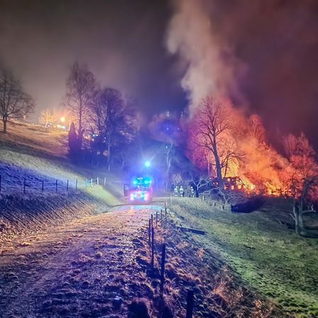 Ein Feuerwehrwagen mit blinkenden blauen Lichtern fährt bei Nacht eine Straße hinunter, mit einem großen Feuer, das am Hang brennt.