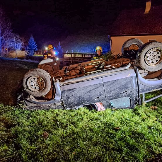 Ein Pickup-Truck ist auf einem Grashang in der Nacht umgekippt. Zwei Feuerwehrleute in gelben Helmen stehen in der Nähe, einer hält eine Taschenlampe.