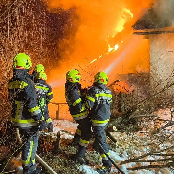Feuerwehrleute in Schutzkleidung bekämpfen ein Feuer, das ein Haus einnimmt, wobei einer Wasser spritzt und andere mit Schläuchen bereitstehen.