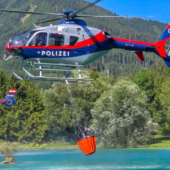 Ein Polizeihubschrauber lässt einen roten Rettungskorb in einen See hinab, mit Bergen und Bäumen im Hintergrund.