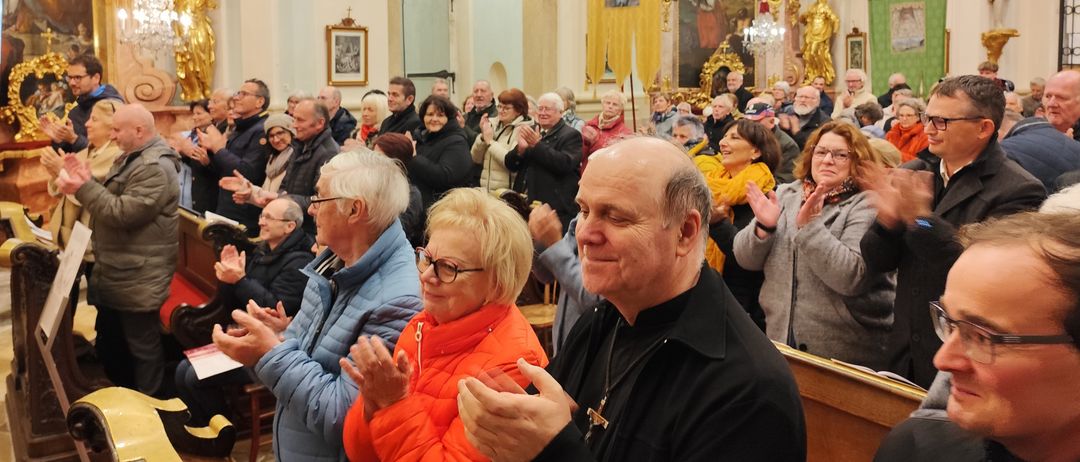 Eine große Gemeinde versammelt sich in einer Kirche, klatscht und lächelt. Die Kirche ist prächtig, mit goldenen Dekorationen und Gemälden an den Wänden. Der Altar ist sichtbar, und ein Priester steht davor.