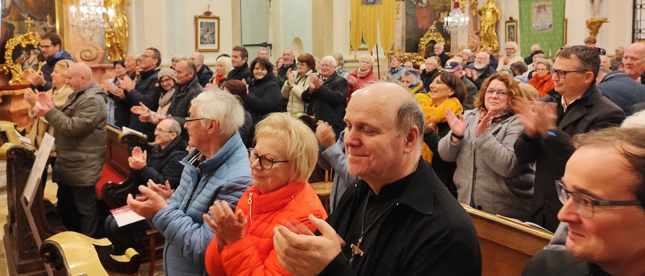 Eine große Gemeinde versammelt sich in einer Kirche, klatscht und lächelt. Die Kirche ist prächtig, mit goldenen Dekorationen und Gemälden an den Wänden. Der Altar ist sichtbar, und ein Priester steht davor.