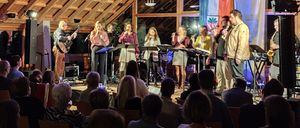 Eine Gruppe Musiker tritt auf einer hell beleuchteten Bühne in einem Raum mit Holzwänden auf, während ein Publikum zuschaut.