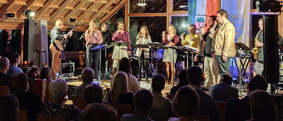 Eine Gruppe Musiker tritt auf einer hell beleuchteten Bühne in einem Raum mit Holzwänden auf, während ein Publikum zuschaut.