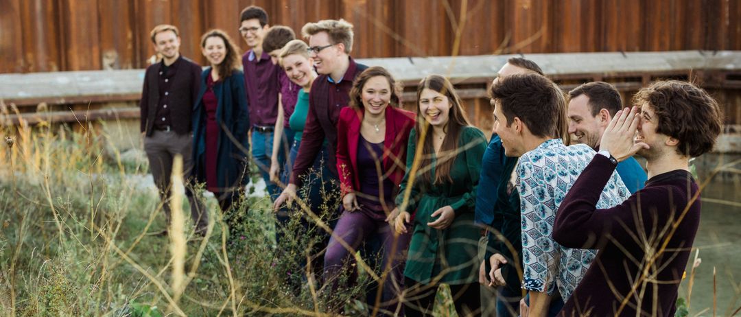 Eine Gruppe von Menschen steht in einem hohen Grasfeld, einige lächeln und lachen. Hinter ihnen ist eine Holzstruktur zu sehen.