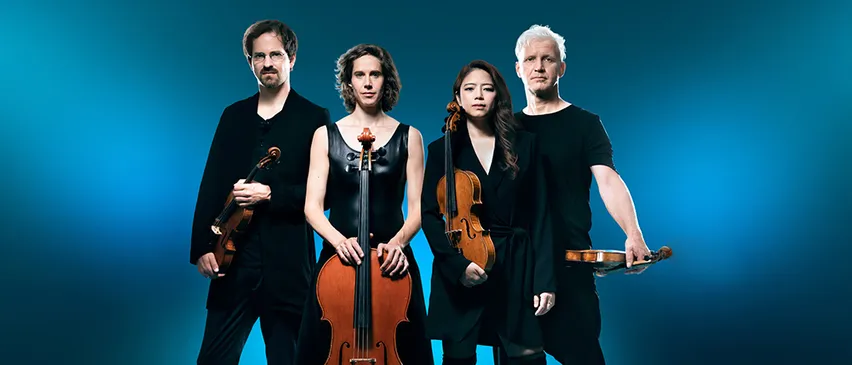 Vier Musiker stehen zusammen, jeder hält eine Geige oder ein Cello. Zwei Frauen und zwei Männer. Sie sind in Schwarz gekleidet, vor einem blauen Hintergrund.