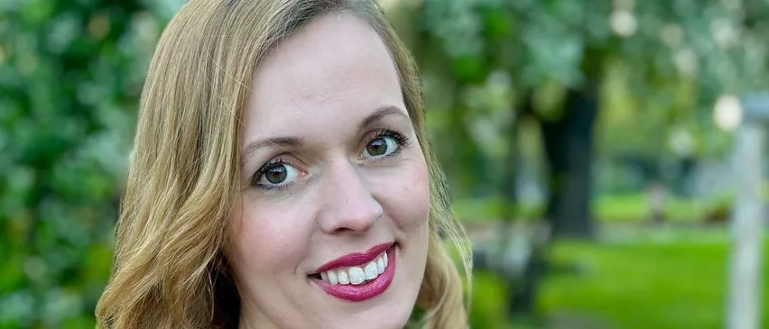 Eine Frau mit blonden Haaren und einem lila Kleid lächelt fröhlich in einem Park mit saftigem grünem Gras.