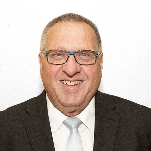 Ein Mann mit Brille und Anzug lächelt in die Kamera. Er hat graues Haar und ein weißes Hemd mit einer grauen Krawatte.