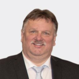 Ein Mann mit einem Schnurrbart und braunem Haar, lächelnd, trägt einen schwarzen Anzug und ein weißes Hemd mit einer hellblauen Krawatte.