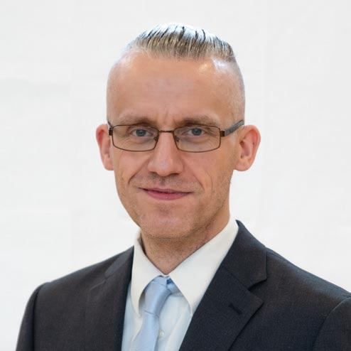 Ein Mann mit kurzen Haaren, der eine Brille, einen Anzug und eine hellblaue Krawatte trägt. Er lächelt mit leicht geöffnetem Mund. Er steht vor einem weißen Hintergrund.