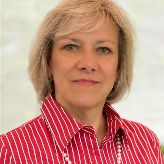Eine Frau mit kurzen blonden Haaren trägt ein rot-weiß gestreiftes Hemd und eine Perlenkette und lächelt.
