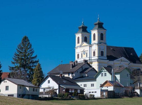 Eine malerische Dorfkirche mit zwei Türmen steht prominent auf einem Hügel, umgeben von einer Ansammlung kleiner, charmanter Häuser unter einem klaren blauen Himmel.