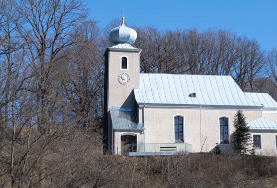 Eine weiße Kirche mit einer Kuppel und einem Glockenturm steht auf einem Hügel, umgeben von Bäumen im Winter.
