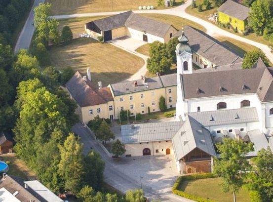 Ein Luftbild eines Klosters mit einem Pool, umgeben von Bäumen und Gebäuden. Das Kloster hat ein großes, graues Dach und einen Glockenturm.
