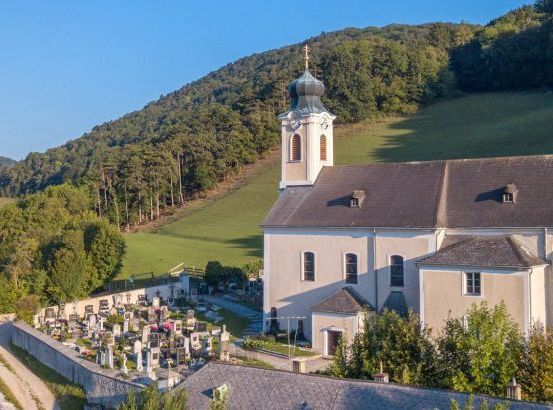 Eine Draufsicht auf eine Kirche mit Turmspitze und Friedhof, umgeben von üppigem Grün und Bergen unter einem klaren Himmel.