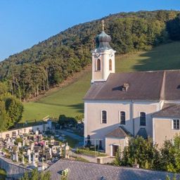 Eine Draufsicht auf eine Kirche mit Turmspitze und Friedhof, umgeben von üppigem Grün und Bergen unter einem klaren Himmel.