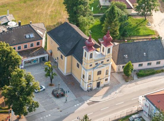 Ein Luftbild eines Dorfes zeigt eine gelbe Kirche mit zwei Türmen. Die Kirche ist von verschiedenen Gebäuden umgeben, einige mit roten Dächern. Bäume und Pflanzen zieren das Gebiet, und eine Straße mit geparkten Autos verläuft durch das Dorf.