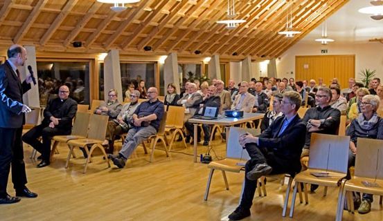 Ein Mann spricht in ein Mikrofon in einem Konferenzsaal und wendet sich an ein Publikum, das in Stühlen sitzt. Der Raum hat Holzböden und Balken, mit einem Projektor auf einem Tisch. Die Leute hören aufmerksam zu.