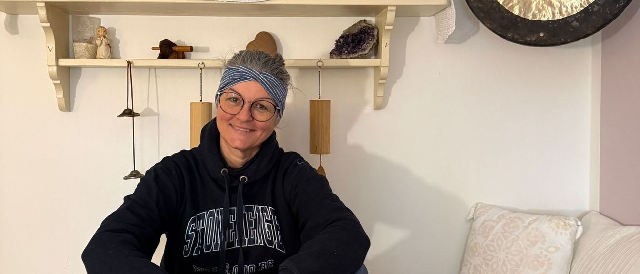 Eine Frau in einem Hoodie sitzt lächelnd auf einem Bett, mit Singschalen und einem Gong an der Wand.