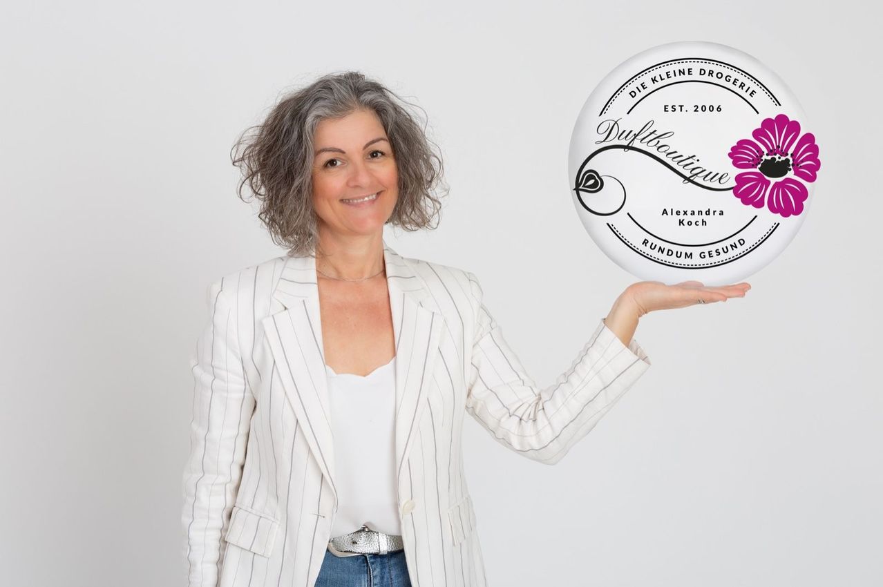 Eine Frau mit grauem Haar und einem weißen Blazer hält ein rundes Produktetikett von Duftboutique Alexandra Koch. Das Etikett zeigt eine rosa Blume und Text auf Deutsch.