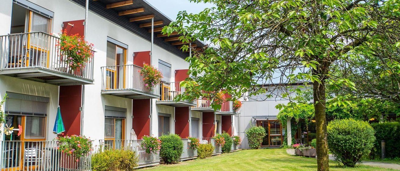Außenansicht eines modernen Gebäudes mit roten und weißen Wänden, mehreren Balkonen, die mit leuchtenden Blumen geschmückt sind, und einem gepflegten Garten.