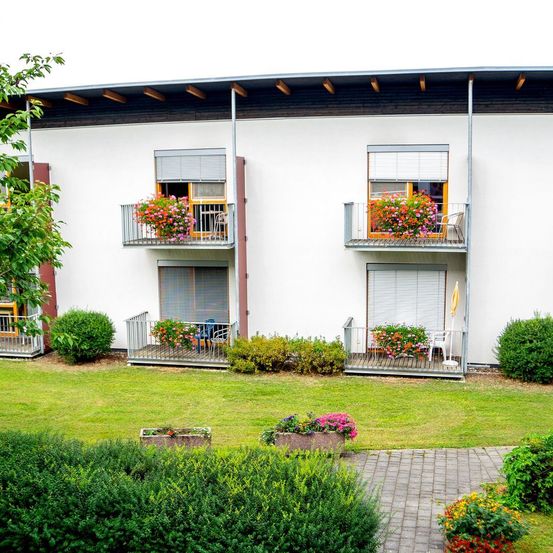 Ein weißes Gebäude mit mehreren Balkonen, die mit Blumen geschmückt sind, einem Rasenbereich und verschiedenen Sträuchern und Pflanzen. Die Balkone haben Stühle und Tische.