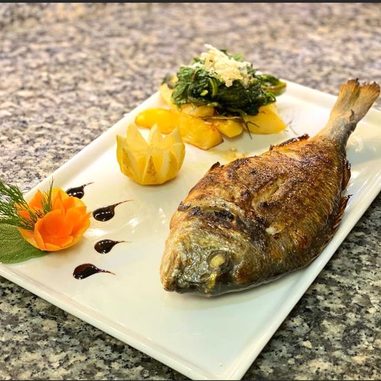 Ein ganzer gebratener Fisch auf einem weißen Teller mit einer Zitrone, Kräutern und Gemüse. Der Fisch ist goldbraun mit einer knusprigen Haut.