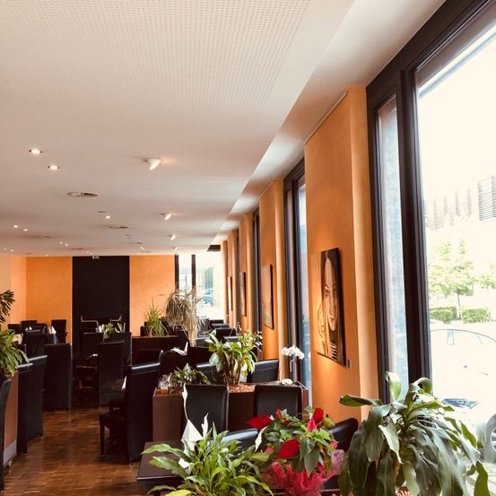 Ein Restaurant mit Holzböden und beigefarbenen Wänden, voller Tische und Stühle. Die Decke ist weiß, und die Fenster sind groß mit schwarzen Rahmen. Es gibt Pflanzen und Gemälde an den Wänden.