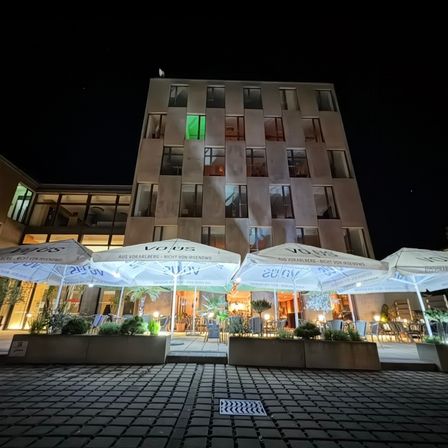 Ein Gebäude mit vielen Fenstern und Lichtern bei Nacht. Ein Restaurantbereich mit Sonnenschirmen und Pflanzen draußen.