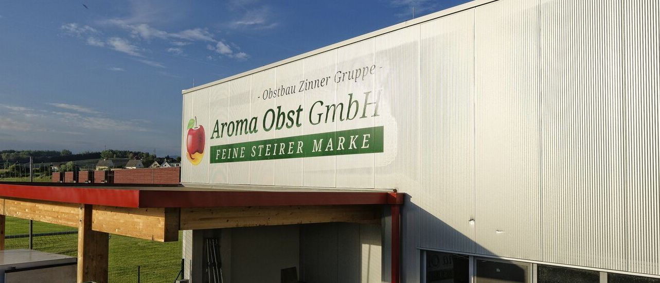 Das Äußere eines weißen Gebäudes mit einem roten Dach und einem großen Schild, das 'Aroma Obst GmbH Feine Steirer Marke' liest. Stapel von grünen Kisten sind auf der linken Seite sichtbar.