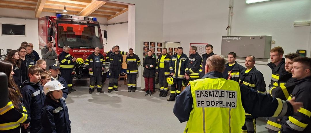 Eine Gruppe von Feuerwehrleuten steht um einen Feuerwehrwagen, einige tragen reflektierende Westen. Ein Mann in einer gelben Weste spricht zu ihnen. Ein Kind mit weißem Helm steht in der Nähe.