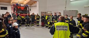 Eine Gruppe von Feuerwehrleuten steht um einen Feuerwehrwagen, einige tragen reflektierende Westen. Ein Mann in einer gelben Weste spricht zu ihnen. Ein Kind mit weißem Helm steht in der Nähe.