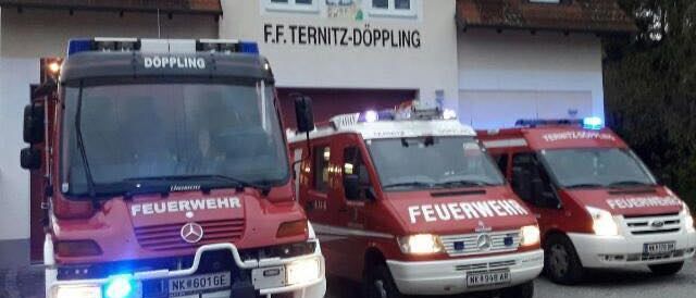 Drei Feuerwehrfahrzeuge sind vor der F.F. Ternitz-Doppling Feuerwehrstation geparkt. Die Fahrzeuge sind rot und weiß mit blinkenden Lichtern auf dem Dach.