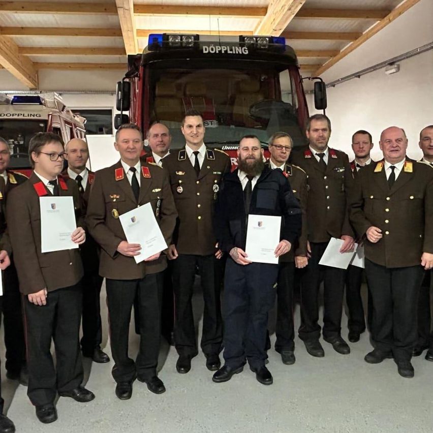 Mehrere Männer in Uniform stehen in einer Gruppe vor einem Feuerwehrauto. Jeder von ihnen hält ein Zertifikat in der Hand, und sie scheinen eine bedeutende Leistung zu feiern.