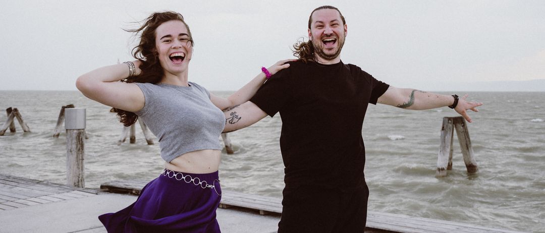 Ein Mann und eine Frau stehen auf einem Pier am Meer und posieren für ein Foto. Die Frau trägt ein graues Oberteil und eine lila Shorts, der Mann ein schwarzes Shirt und schwarze Hose.