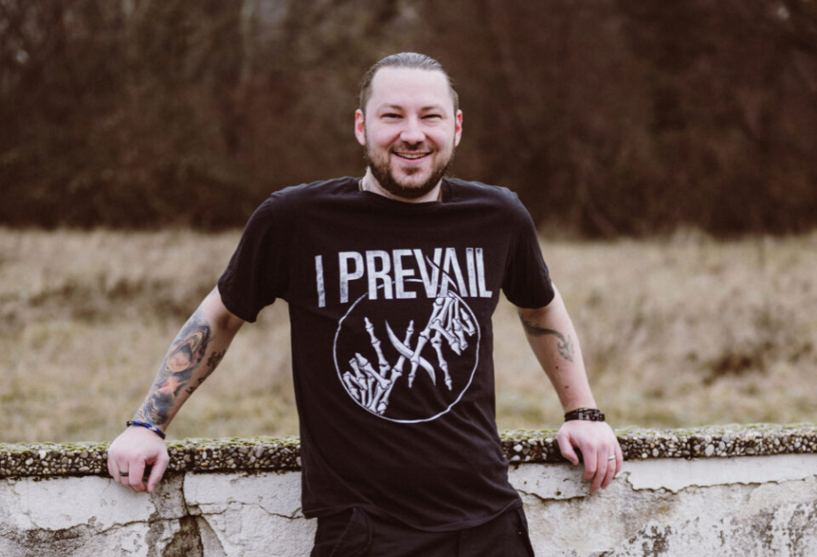Ein Mann mit Tätowierungen an den Armen trägt ein schwarzes T-Shirt mit den Worten 'I PREVAIL' und einer Grafik einer Hand. Er steht an einer Steinmauer.