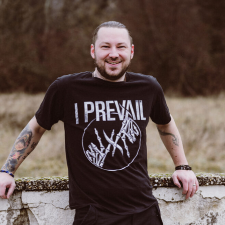 Ein Mann mit Tätowierungen an den Armen trägt ein schwarzes T-Shirt mit den Worten 'I PREVAIL' und einem Skelett-Grafik. Er lächelt und lehnt sich an eine Betonmauer.