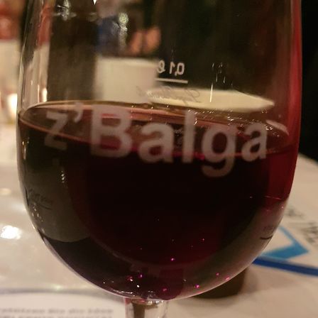 Ein Glas Rotwein mit der Aufschrift Z'Balga steht auf einem Tisch mit einem verschwommenen Hintergrund von sitzenden und trinkenden Personen.