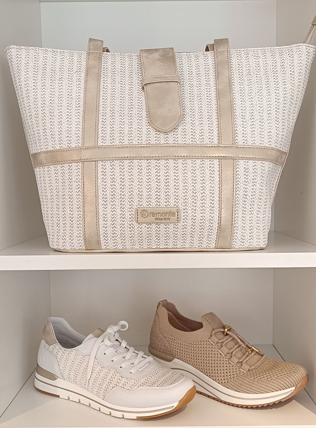 Eine weiße gewebte Tragetasche mit beigefarbenen Akzenten und einem goldenen Logo steht auf einem Regal über einem Paar beigefarbener und weißer Schuhe.
