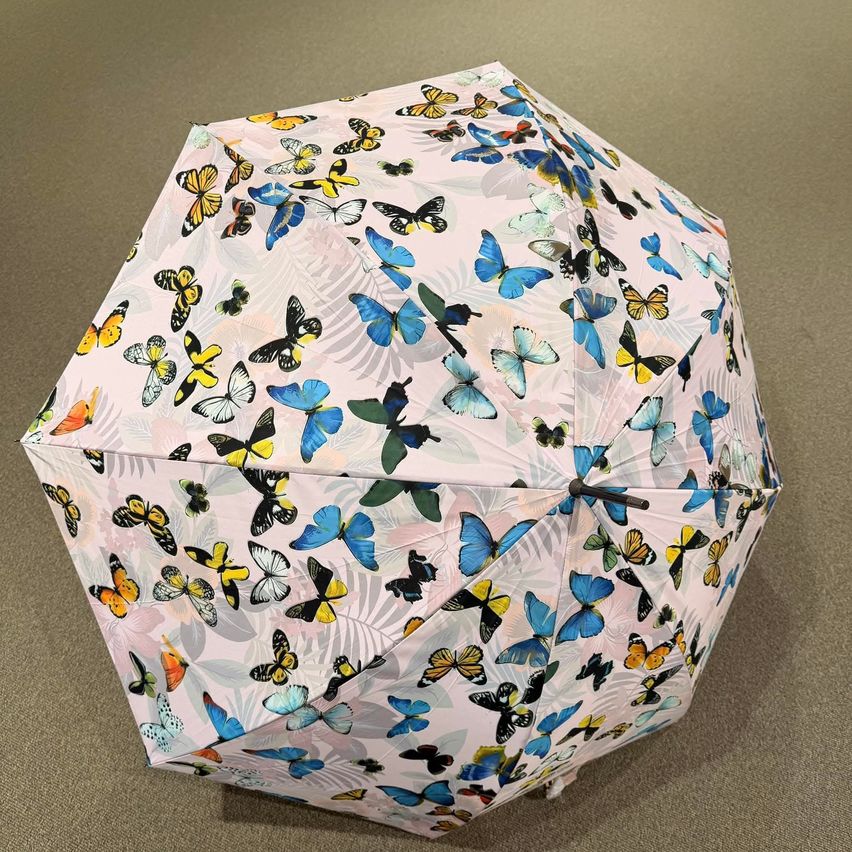 Ein rosafarbener Regenschirm mit mehreren Schmetterlingen in verschiedenen Farben und Mustern, zusammengeklappt und auf einer beigefarbenen Oberfläche platziert.