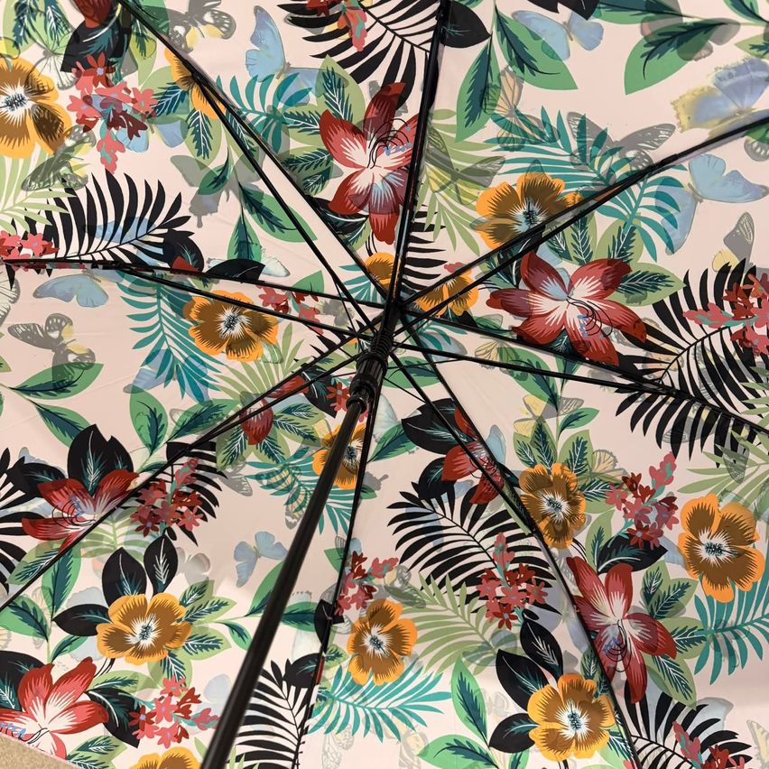 Nahaufnahme eines Regenschirms mit einem lebhaften Blumenmuster, bestehend aus roten, gelben und grünen Blumen und tropischen Blättern. Das Design beinhaltet Schmetterlinge und verschiedene Schattierungen von Rosa und Lila.