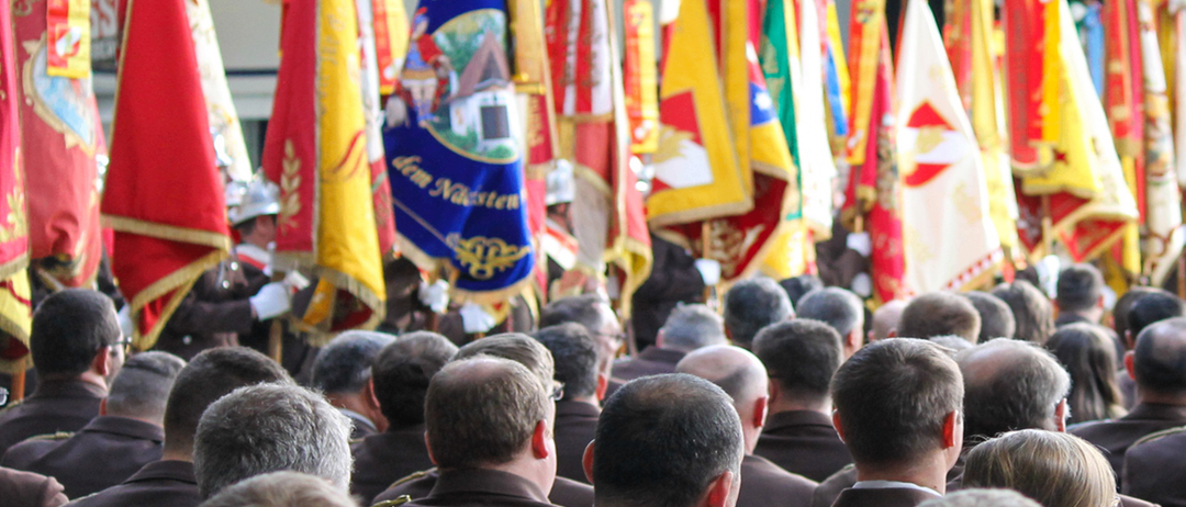 Ein Plakat bewirbt den Tag der Feuerwehr am 29. Mai 2025 um 10:00 Uhr in der Gemeindehalle Wolfau. Viele Menschen sind versammelt und halten verschiedene Fahnen, die wahrscheinlich verschiedene Feuerwehren repräsentieren.