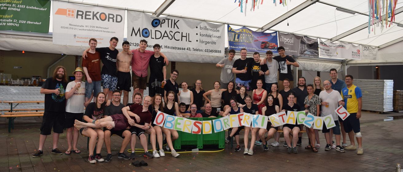 Eine Gruppe von Menschen unter einem Zelt, die lächelnd für ein Foto posiert, einige halten ein Banner mit der Aufschrift 'OBERSDFRFRKITAG202'. Das Zelt hat Banner mit Wörtern wie 'optik', 'moldaschl' und 'rekorD'.