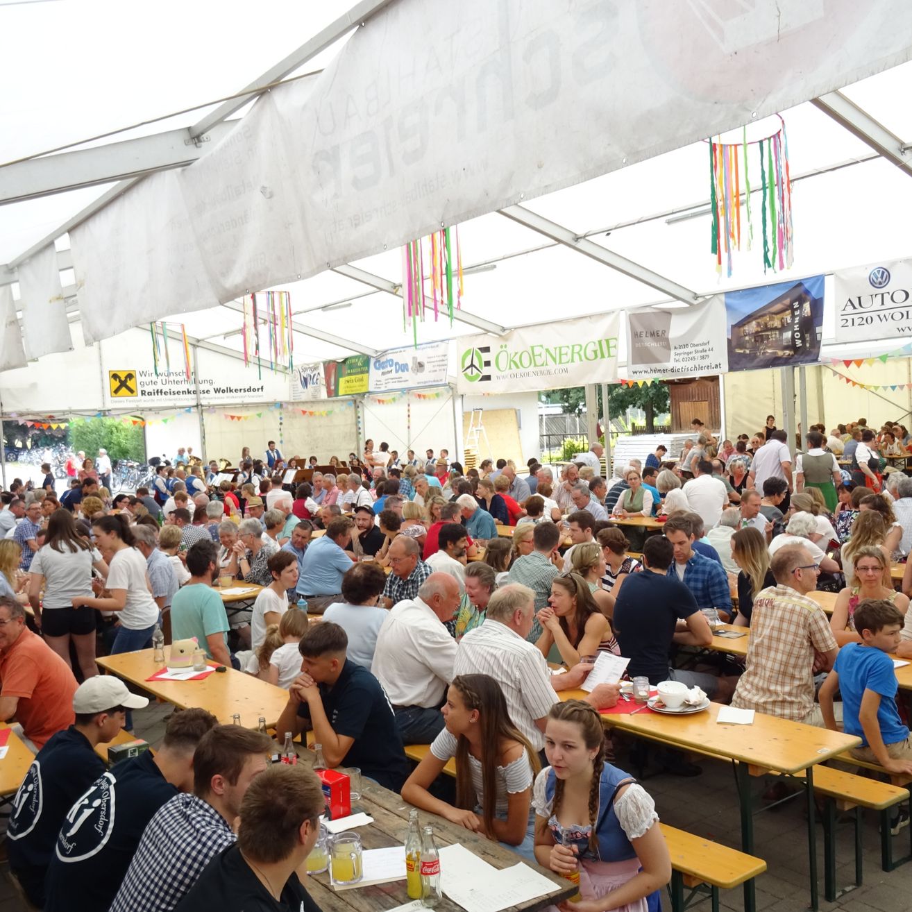 Eine große Gruppe von Menschen sitzt in einem Zeltbereich an Tischen. Viele unterhalten sich, andere essen. Es hängen Schilder und Banner im Zelt.
