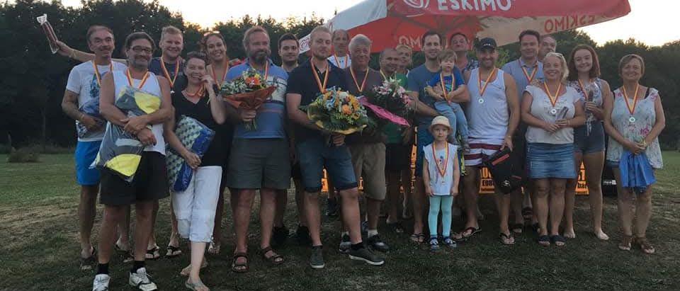 Eine Gruppe von Menschen posiert für ein Foto unter einem Eskimo-Zelt, hält Blumen und Medaillen und steht auf Gras.