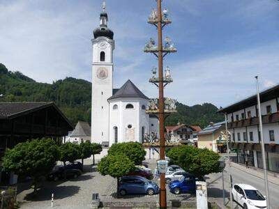 Eine weiße Kirche mit hohem Turm und einer Uhr steht im Zentrum einer Kleinstadt. Autos sind vor der Kirche geparkt. Bäume und ein hoher Pfahl mit Dekorationen umgeben die Kirche. Gebäude säumen die Straße.