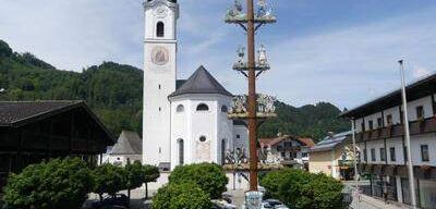 Eine weiße Kirche mit hohem Turm und einer Uhr steht im Zentrum einer Kleinstadt. Autos sind vor der Kirche geparkt. Bäume und ein hoher Pfahl mit Dekorationen umgeben die Kirche. Gebäude säumen die Straße.