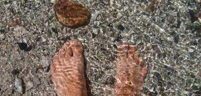 Die Füße einer Person stehen in klarem Wasser mit einem kleinen Stein in der Nähe. Das Wasser ist sauber und flach.